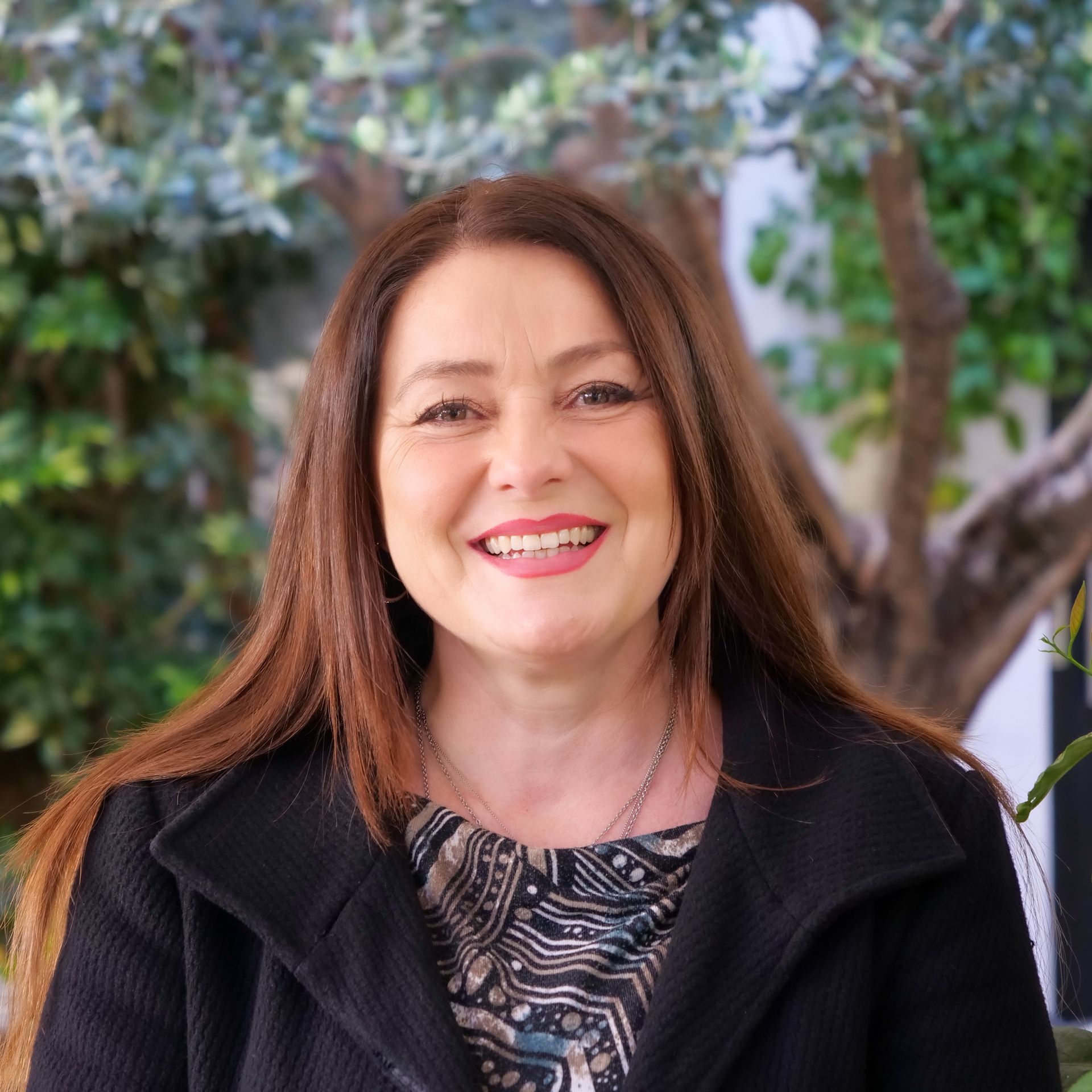 Woman with long brown hair smiles, wearing a black coat and patterned top, outdoors with greenery.