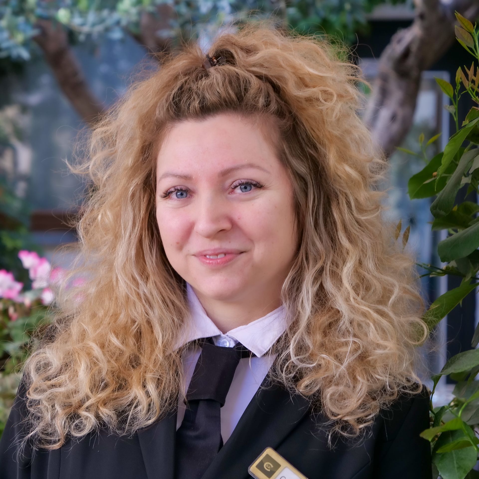 Woman with curly blonde hair in a black suit, smiling, near greenery.