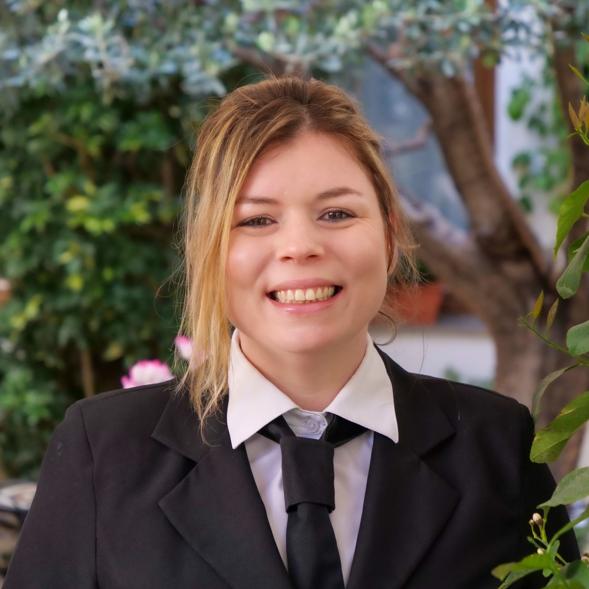 Woman in black suit smiles, standing outdoors near greenery.