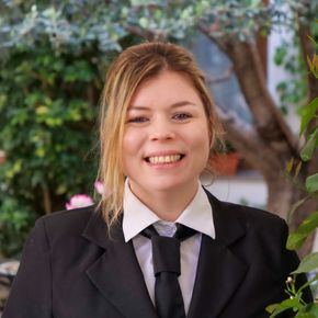 Woman in black suit smiles, standing outdoors near greenery.