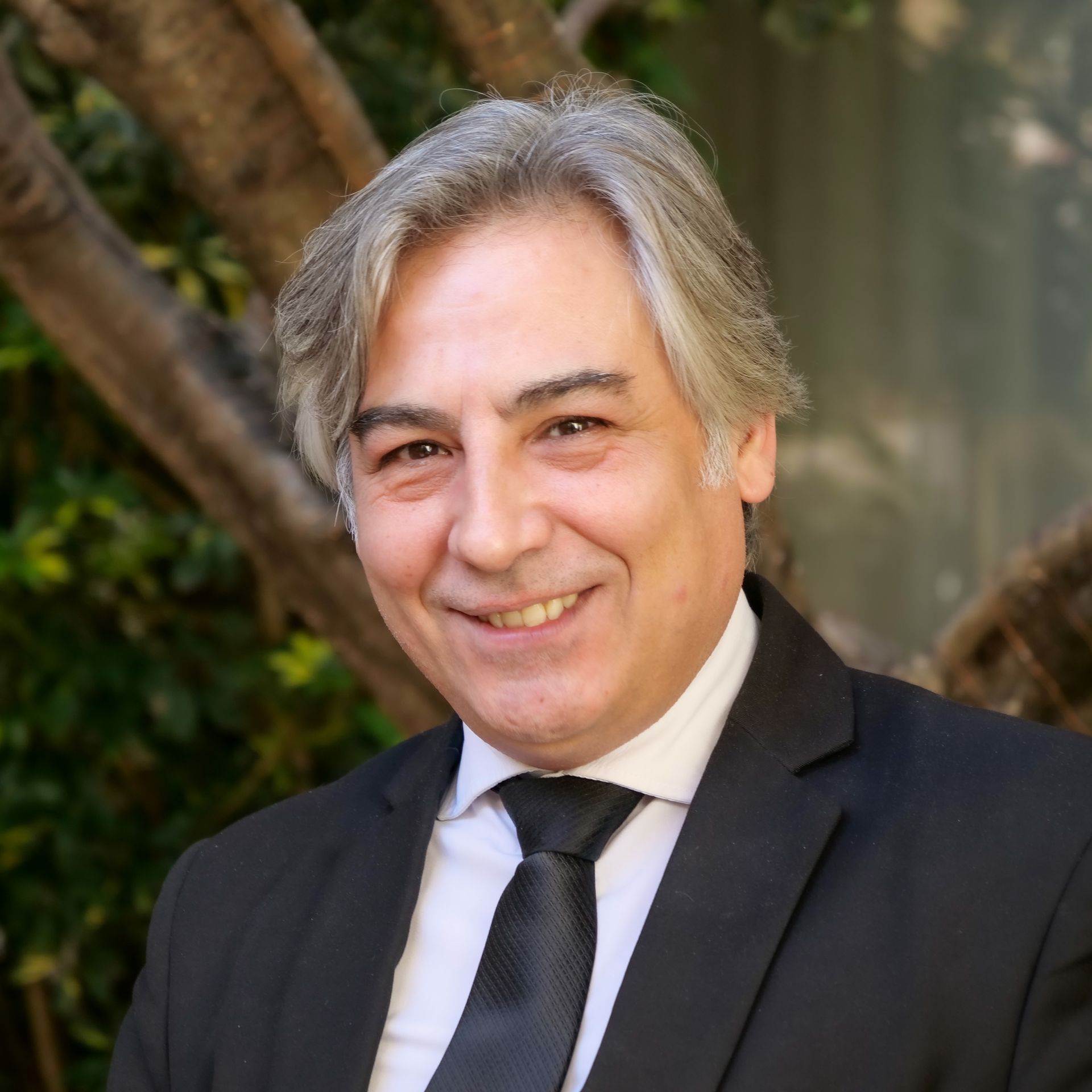 Man in suit smiling, grey hair, outdoors with leafy background.