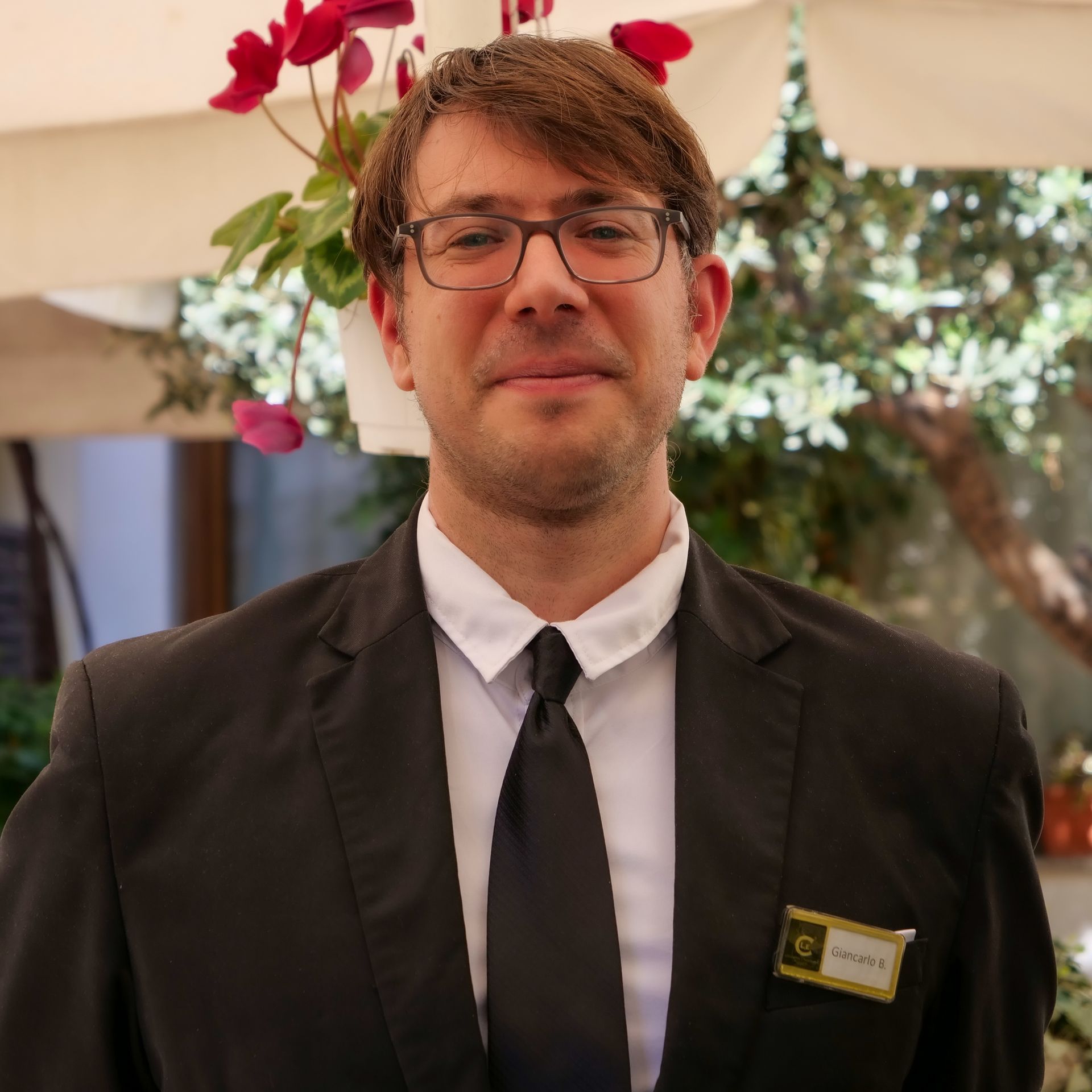 Mann in schwarzem Anzug und Krawatte, mit Brille, lächelt leicht, neben Blumen in einem Innenhof.