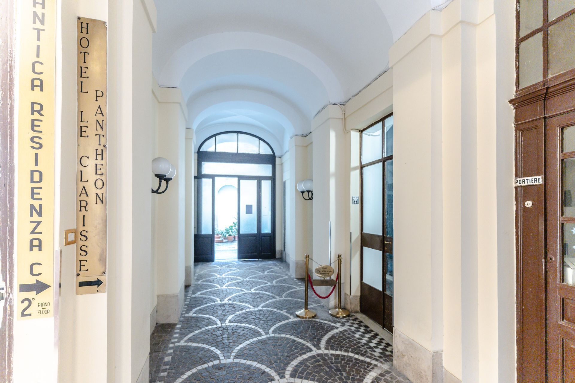 Langer Flur mit gemustertem Steinboden, der zu einer offenen Tür führt, Schilder weisen auf ein Hotel hin.