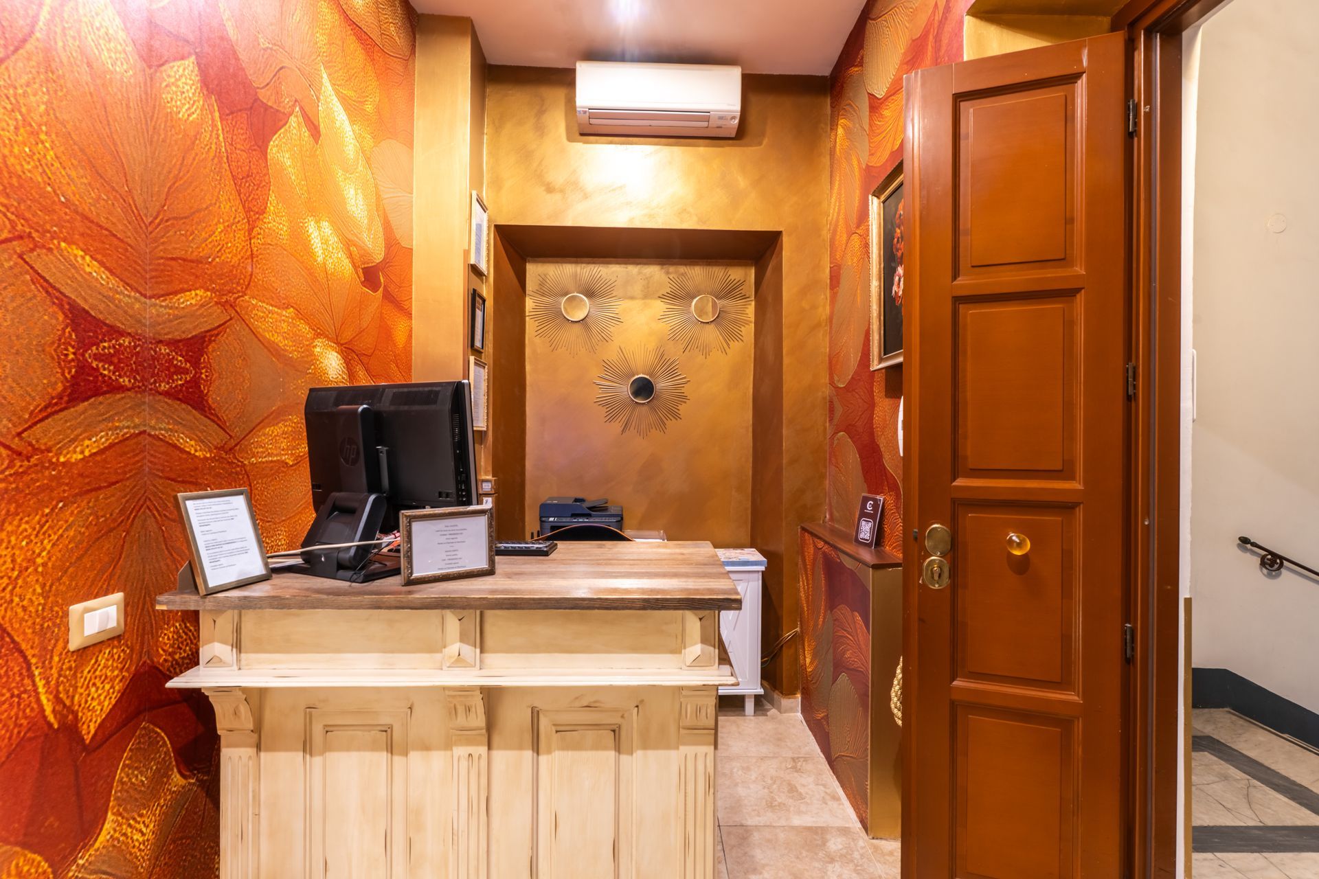 Empfangstresen in einem warm gestalteten Raum; Gold-, Orange- und Brauntöne. Holztür rechts.