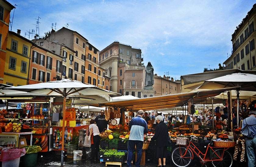 Scène de marché en plein air à Rome : étals, produits frais et passants sous des parasols.