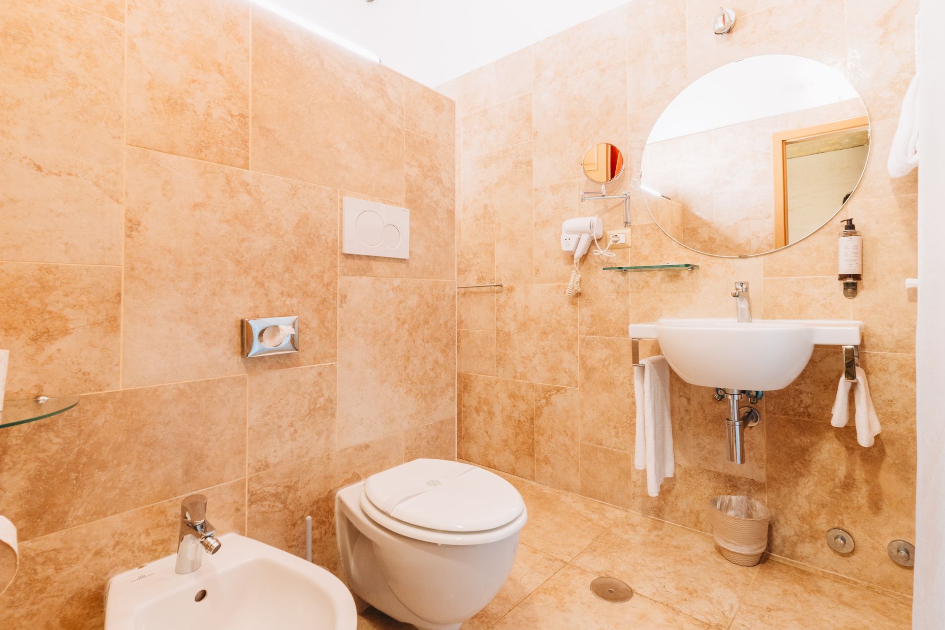 Badezimmer mit beigefarbenen Marmorfliesen, Toilette, Waschbecken, Bidet und rundem Spiegel.