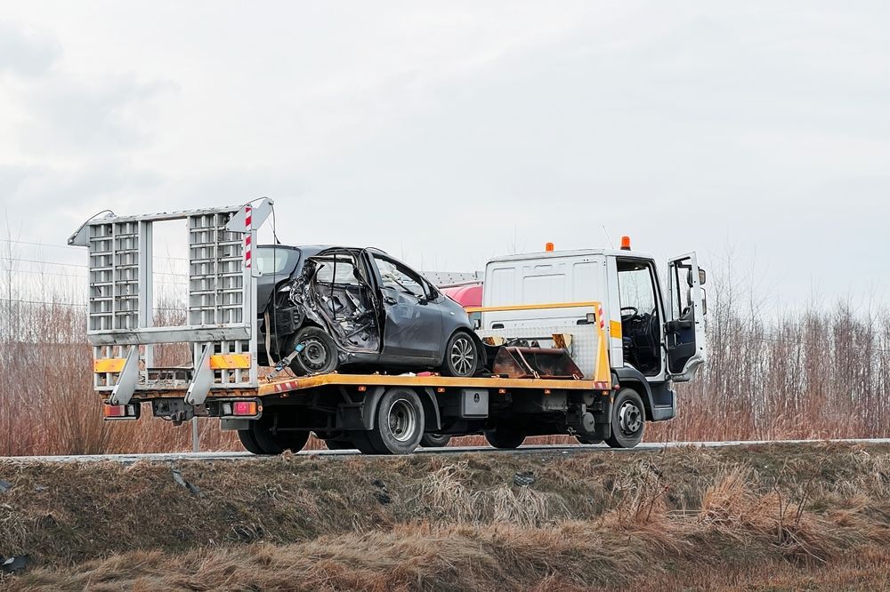 A flatbed tow truck carrying a heavily damaged black car on a road against a backdrop of trees and a cloudy sky.