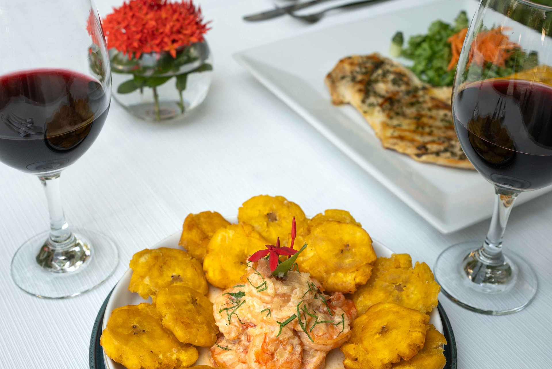 Mesa de cena: plátanos fritos, camarones, pescado a la parrilla, copas de vino tinto y flores.