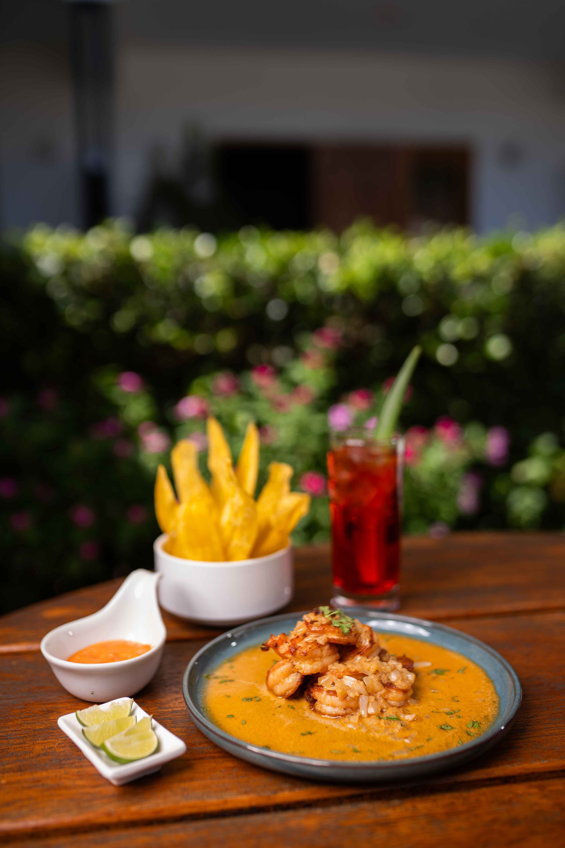 Plato de camarones en salsa de naranja en un plato, con papas fritas, salsa, limón y una bebida en una mesa al aire libre.