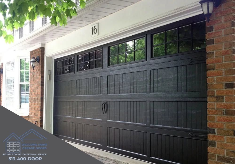 Black garage door with windows on a brick house, framed by white trim and green tree leaves above.