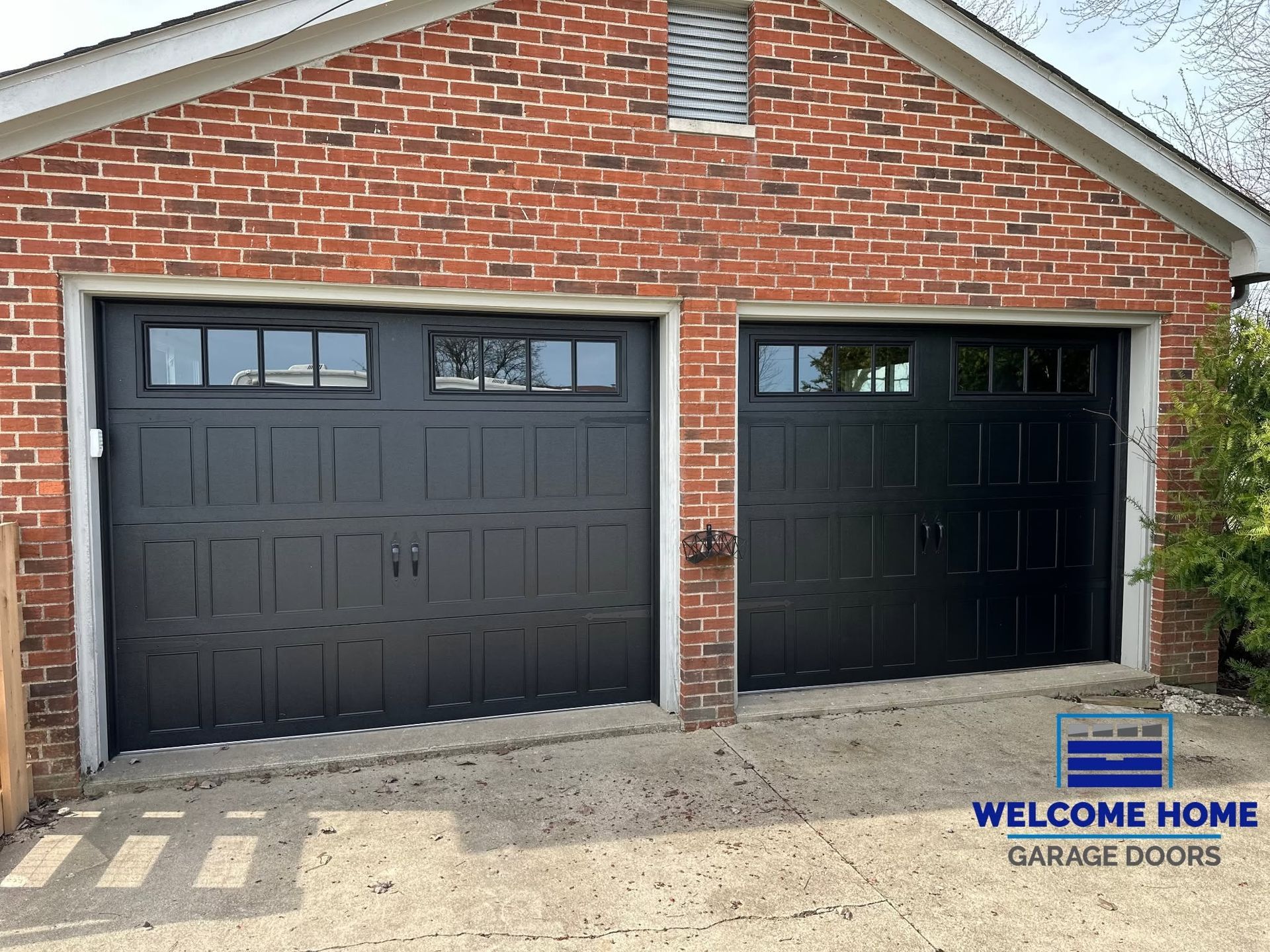 Two black garage doors on a brick house facade, with a welcome mat on the driveway.