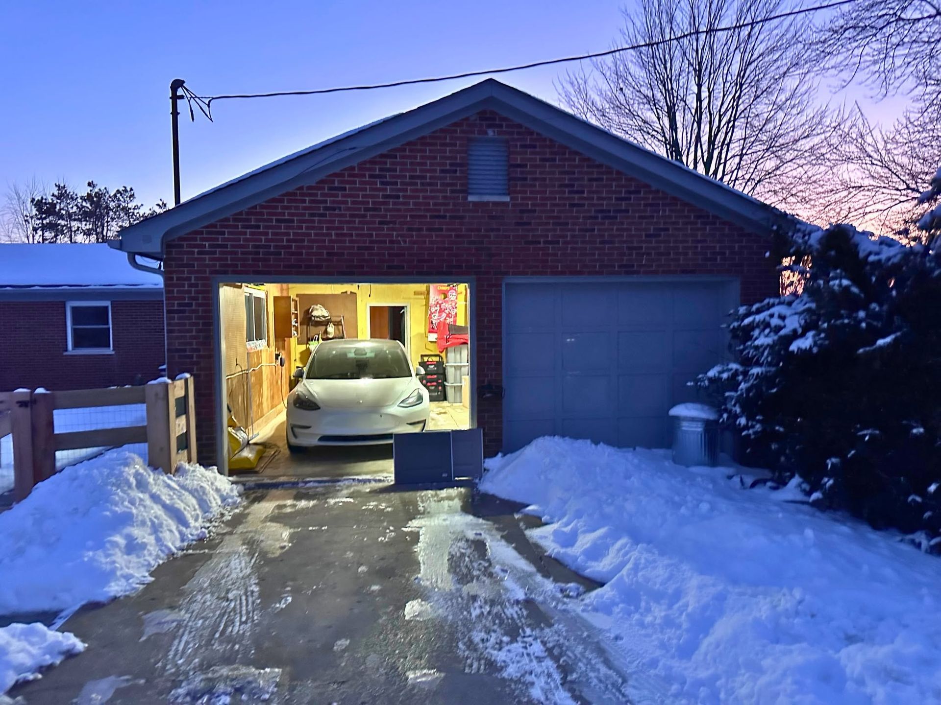 Snowy driveway to a garage with a white car inside, lights on at dusk.