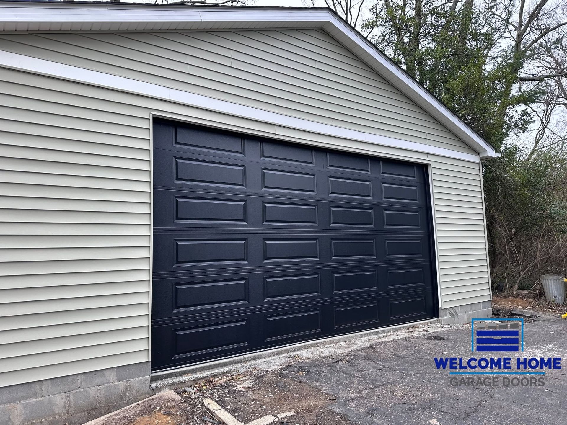 Black garage door on beige-sided house with a gravel driveway and a small “Welcome Home” sign.