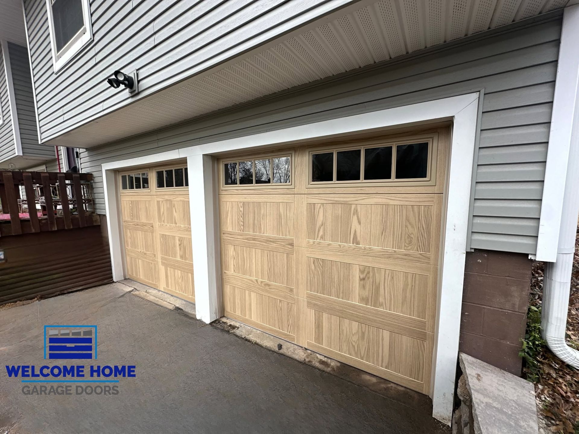 Two unfinished wooden garage doors on a gray house with a small “Welcome Home” sign
