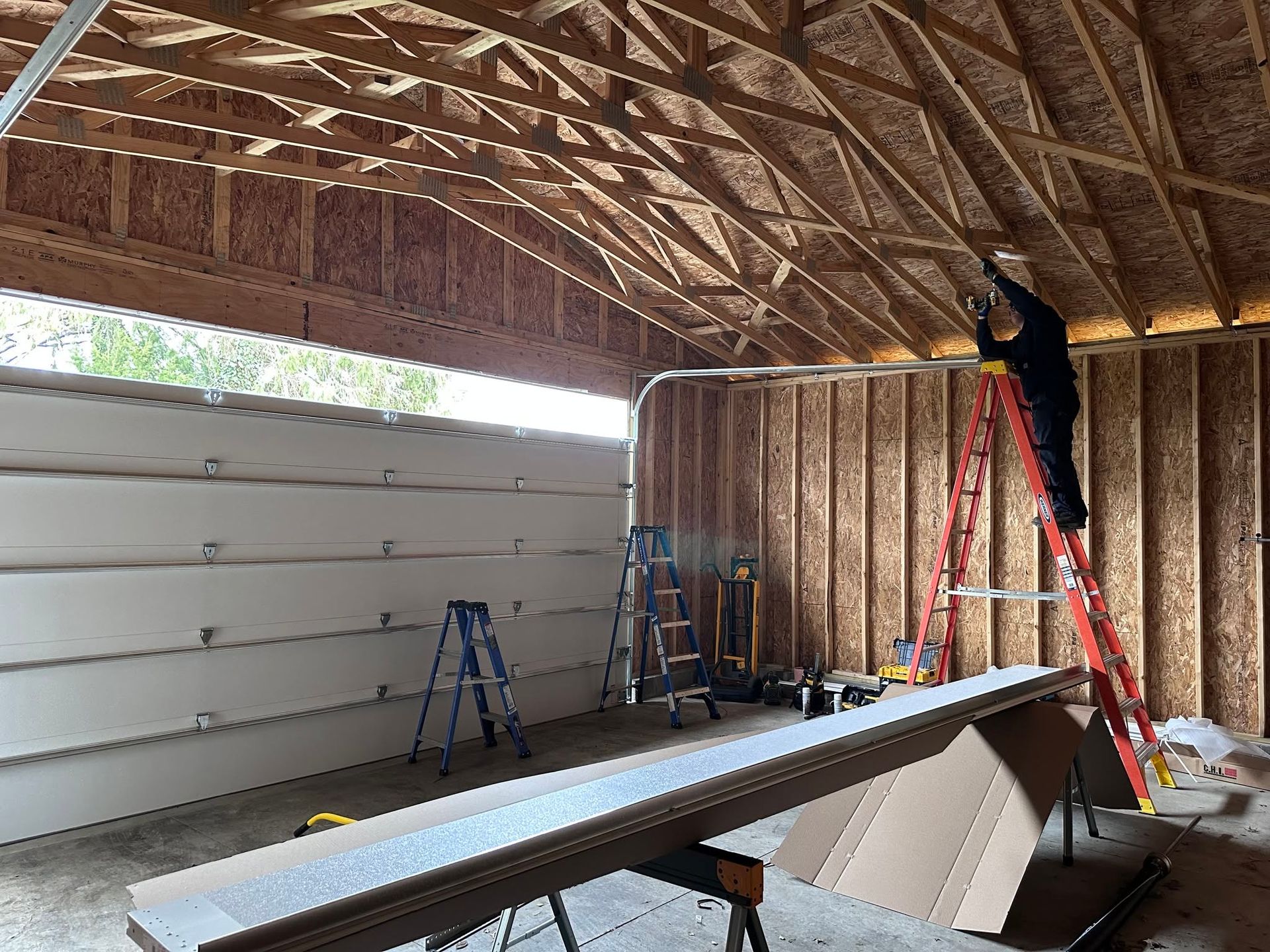Worker on ladder installing insulation in a wood-framed garage with exposed roof trusses.
