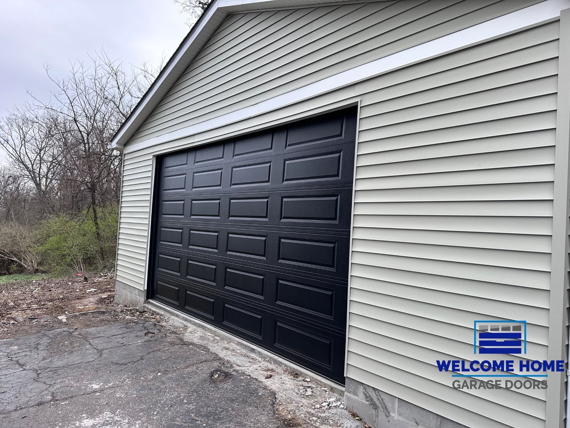 Black garage door on a white-sided building with a small welcome home sign near the driveway.