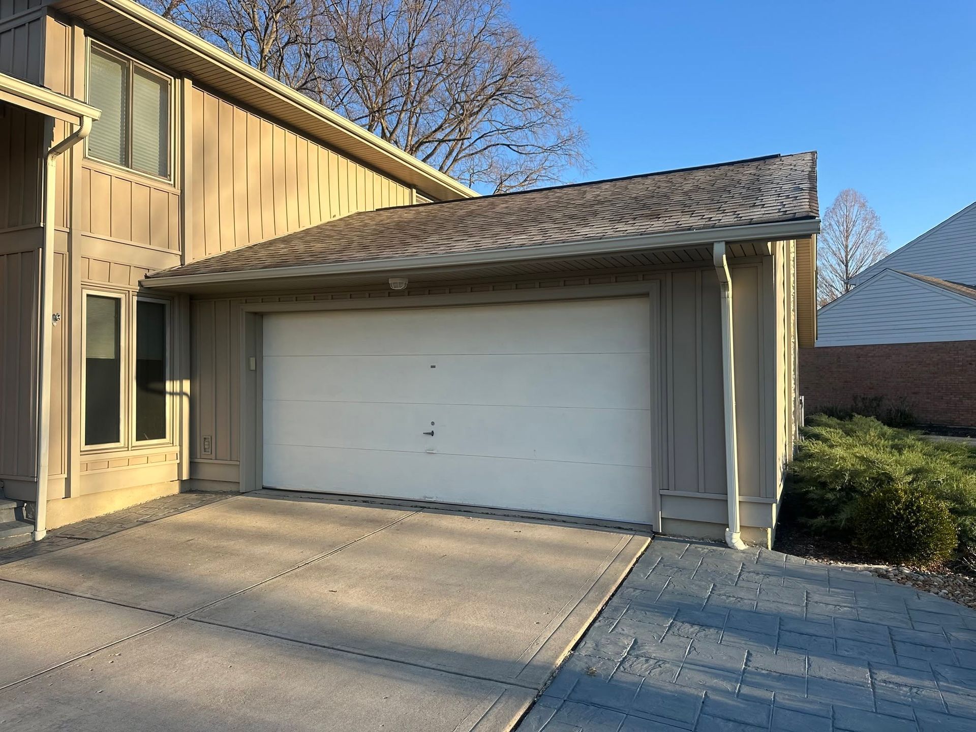 Residential garage with a white door, tan siding, and a paved driveway in sunlight