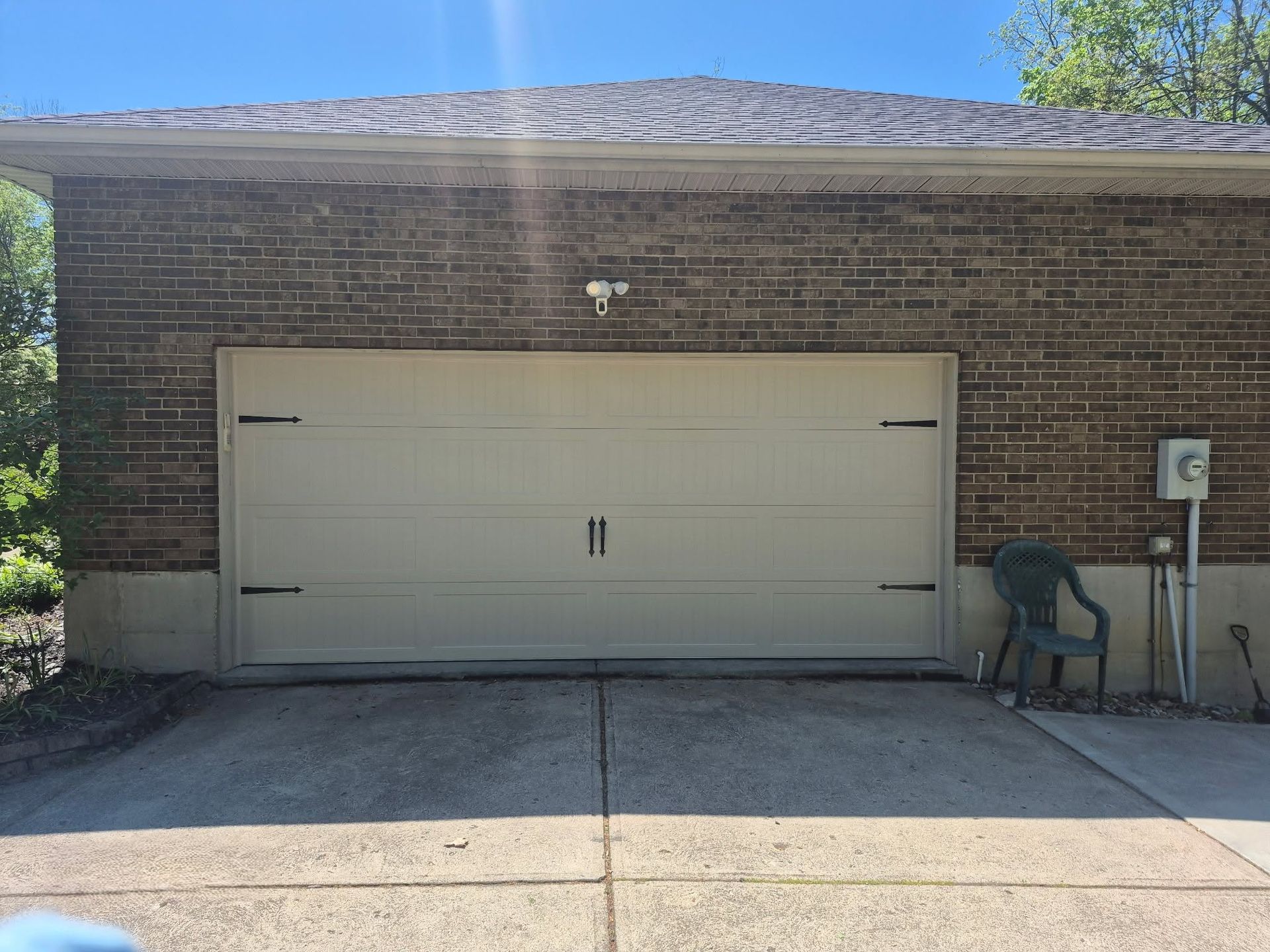 Beige double garage door on a brick house with a concrete driveway and a chair by the side wall