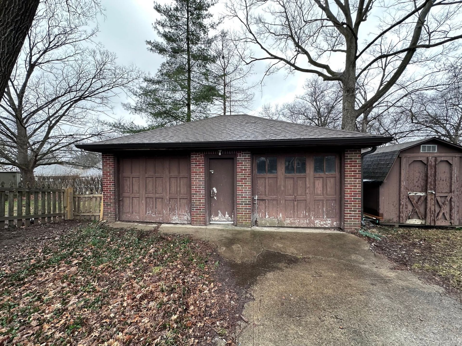 Brick garage with two doors, a leaf-covered yard, and trees in the background