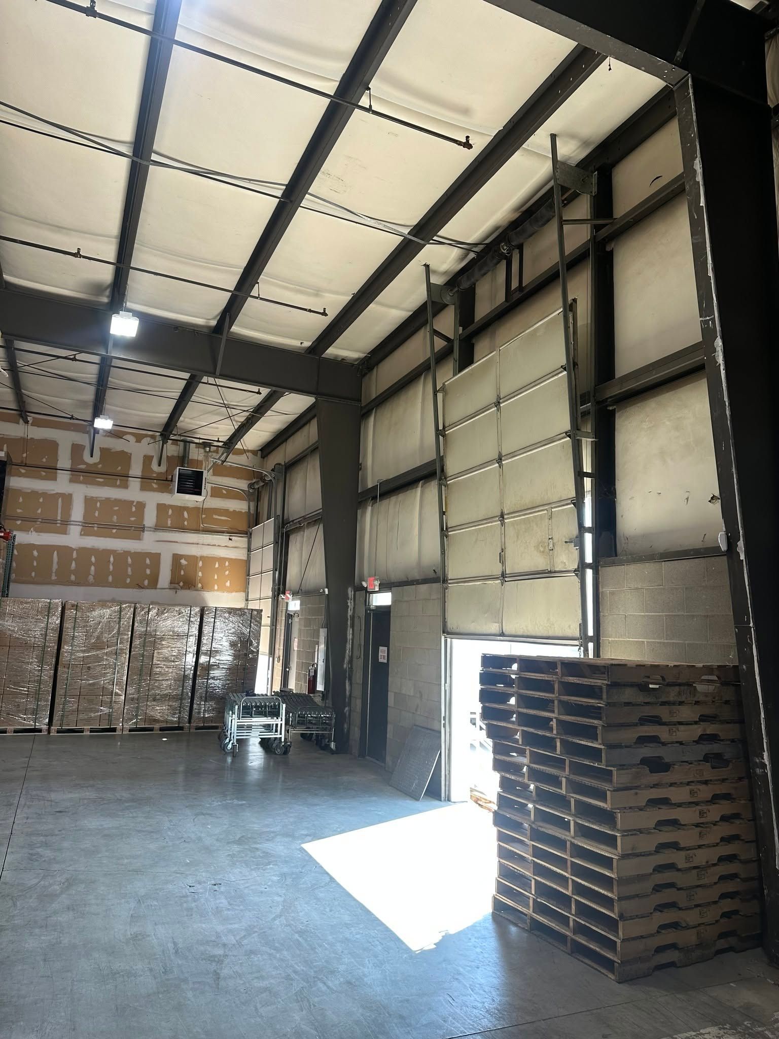 Empty warehouse interior with tall roll-up doors, stacked pallets, and bright sunlight on the floor