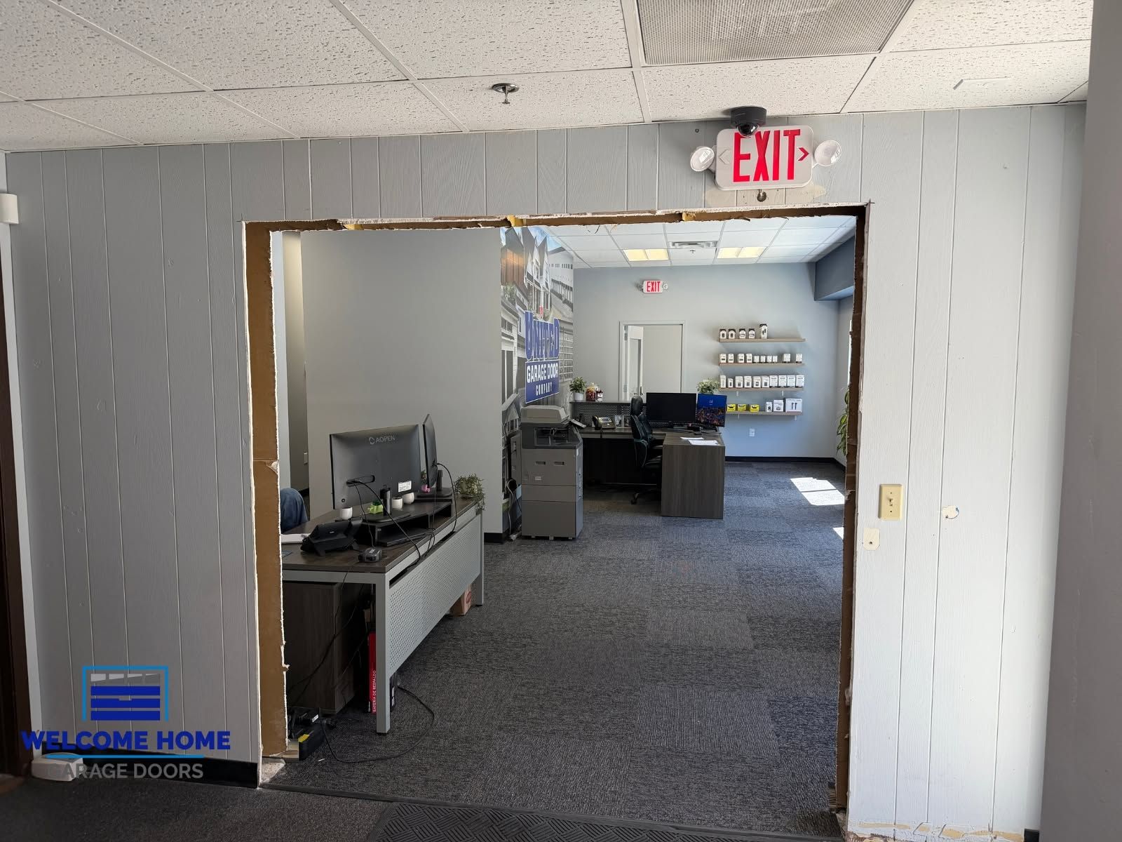 Office hallway leading to a workspace with desks, carpeted floor, and EXIT sign above the doorway