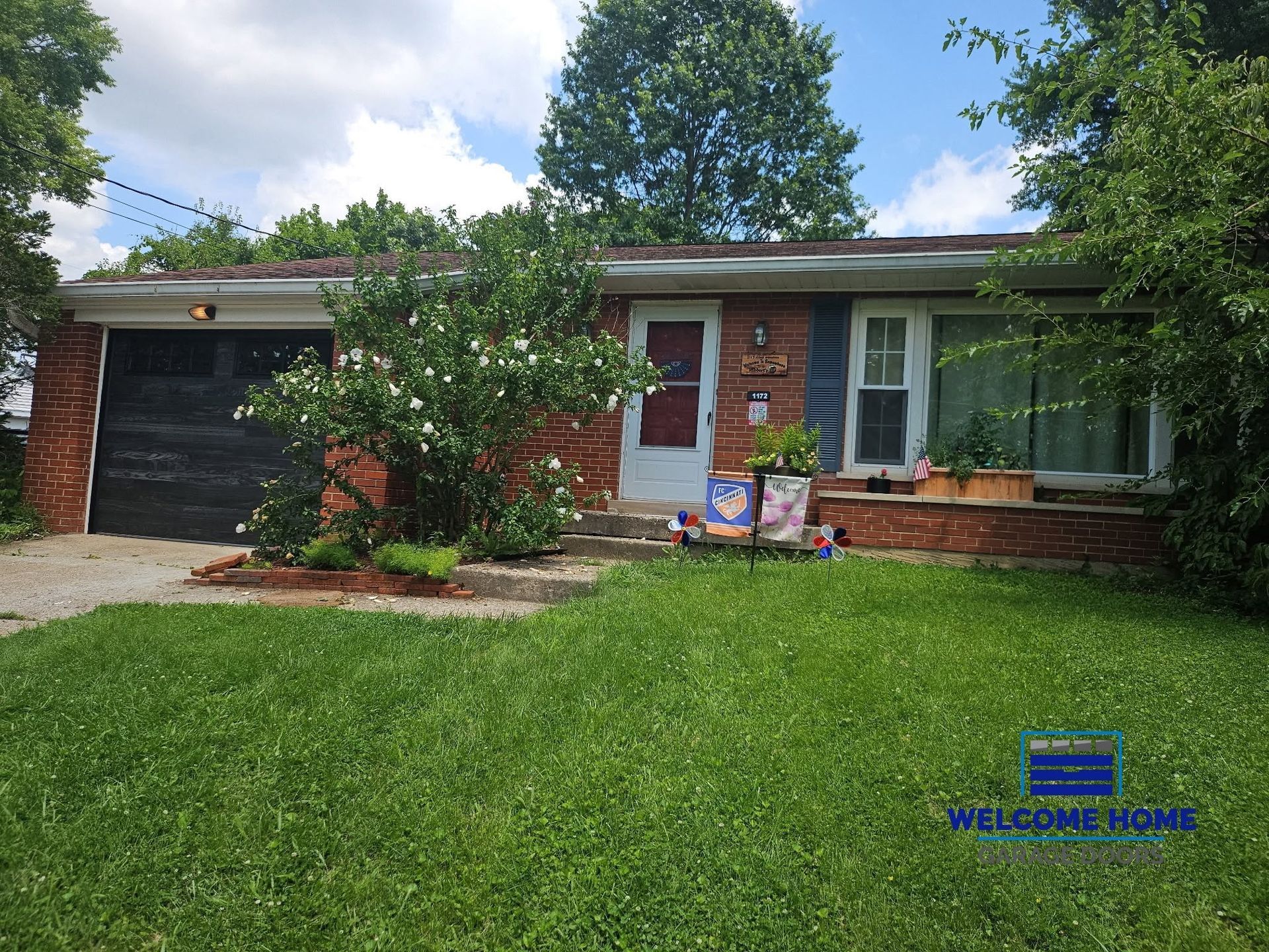 Single-story brick house with black garage door, green lawn, and front porch with potted plants.