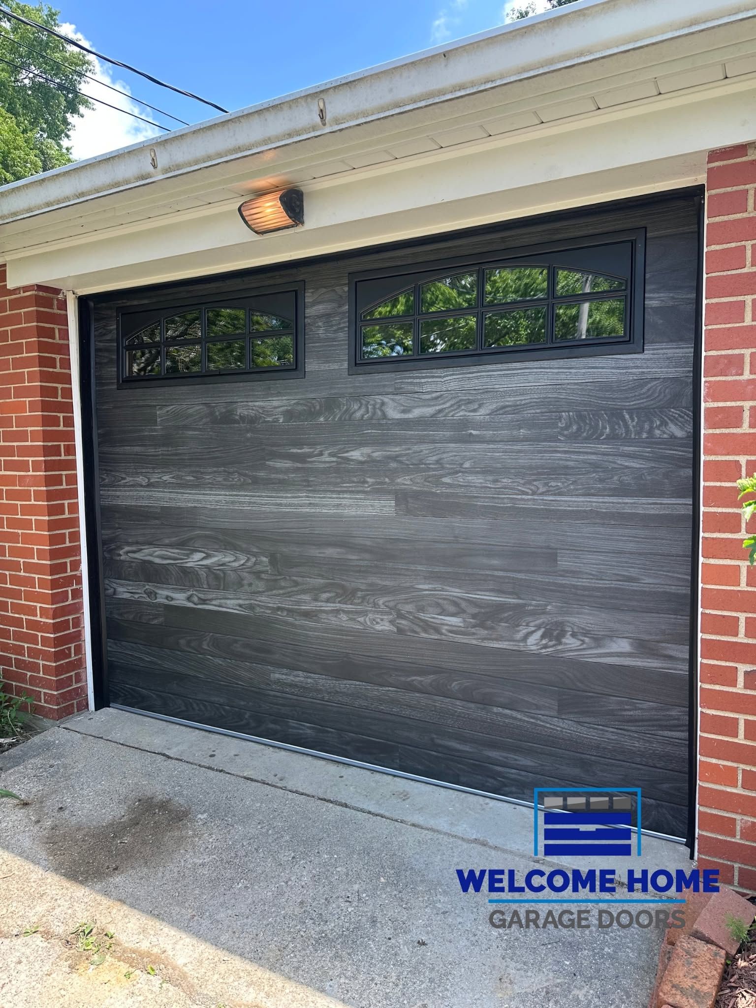 Dark gray garage door with black windows under a white eave, framed by brick walls.