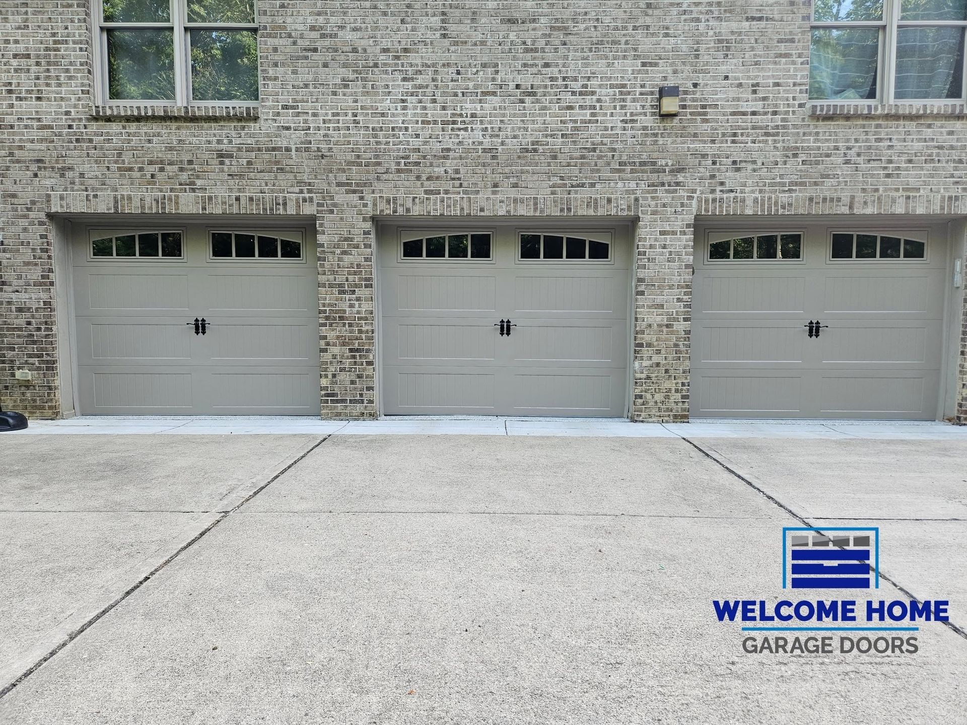 Three beige garage doors on a stone apartment building with a concrete driveway and a Welcome Home sign