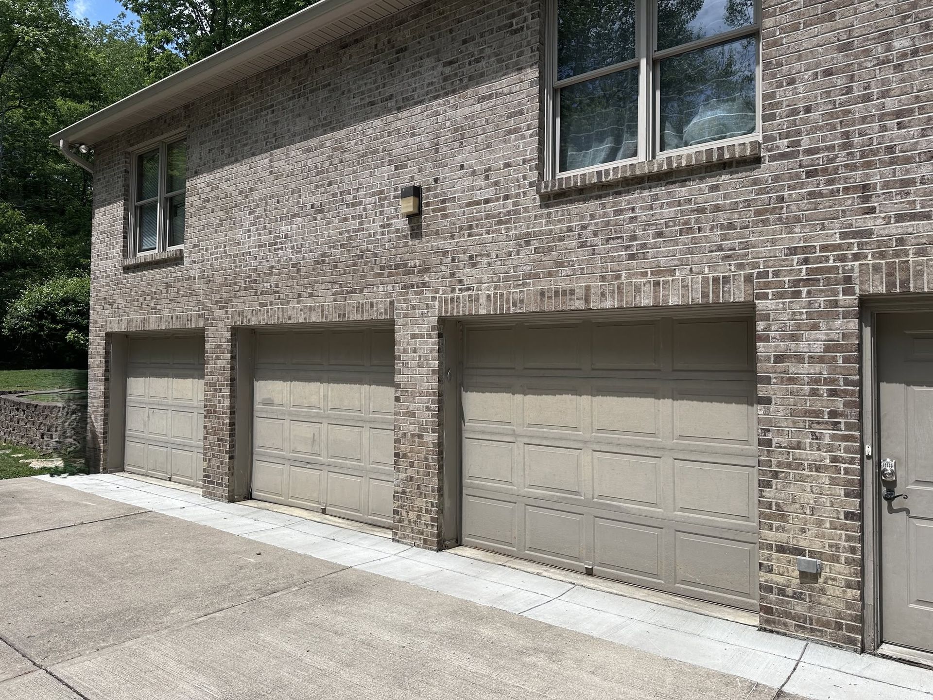Brick apartment garage with three tan roll-up doors and a side entrance, viewed from a driveway.