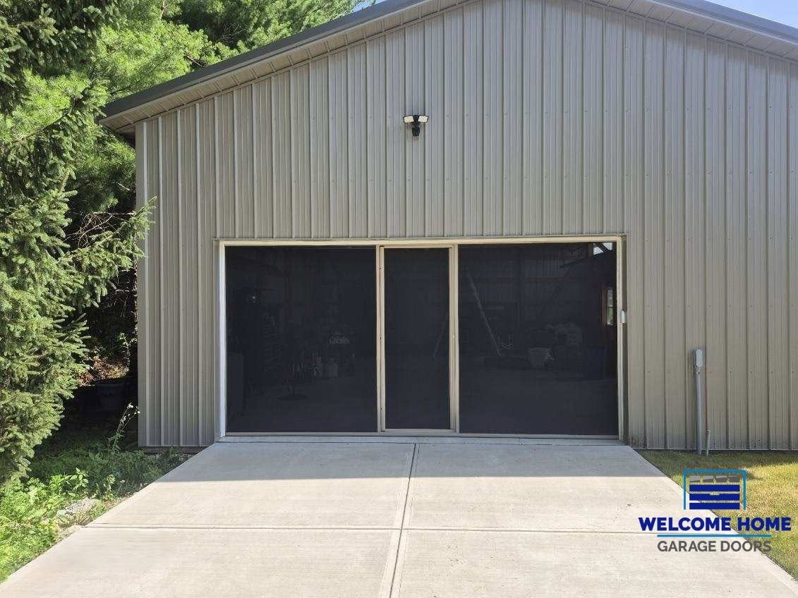 Gray metal building with a large open garage door and concrete driveway under trees.