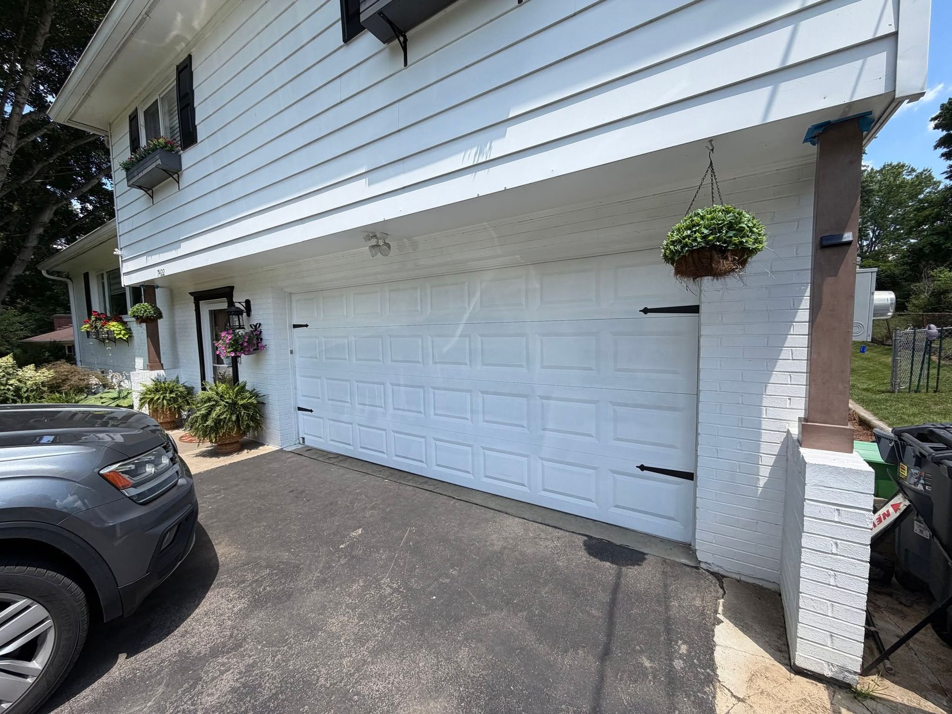 White garage door on a house exterior with a parked dark SUV and hanging flower baskets alongside the driveway
