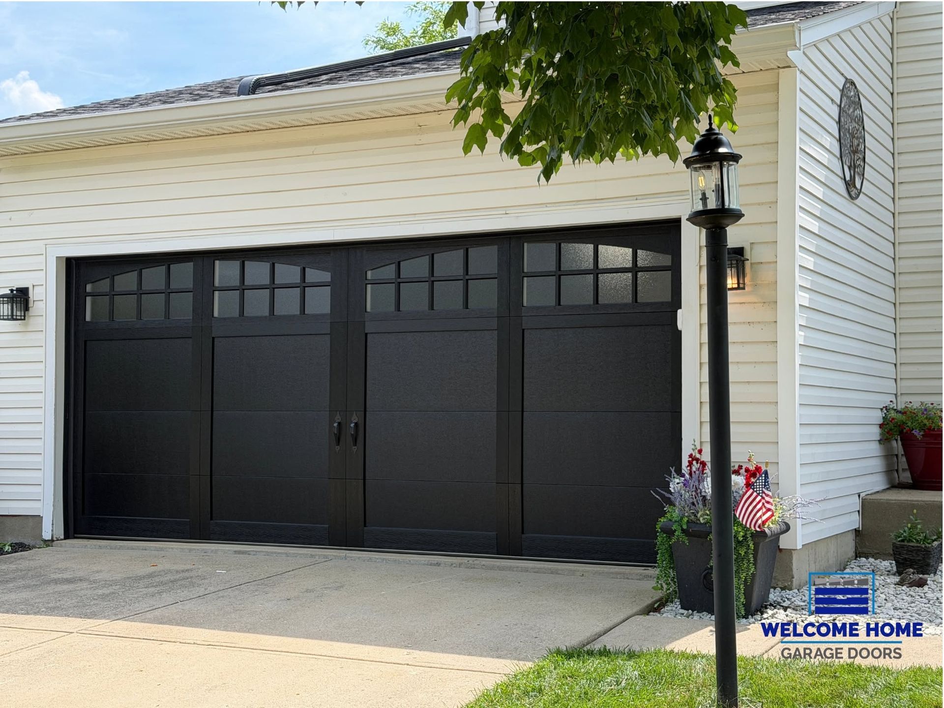 White house exterior with a black double garage door and a small “welcome” sign by the driveway