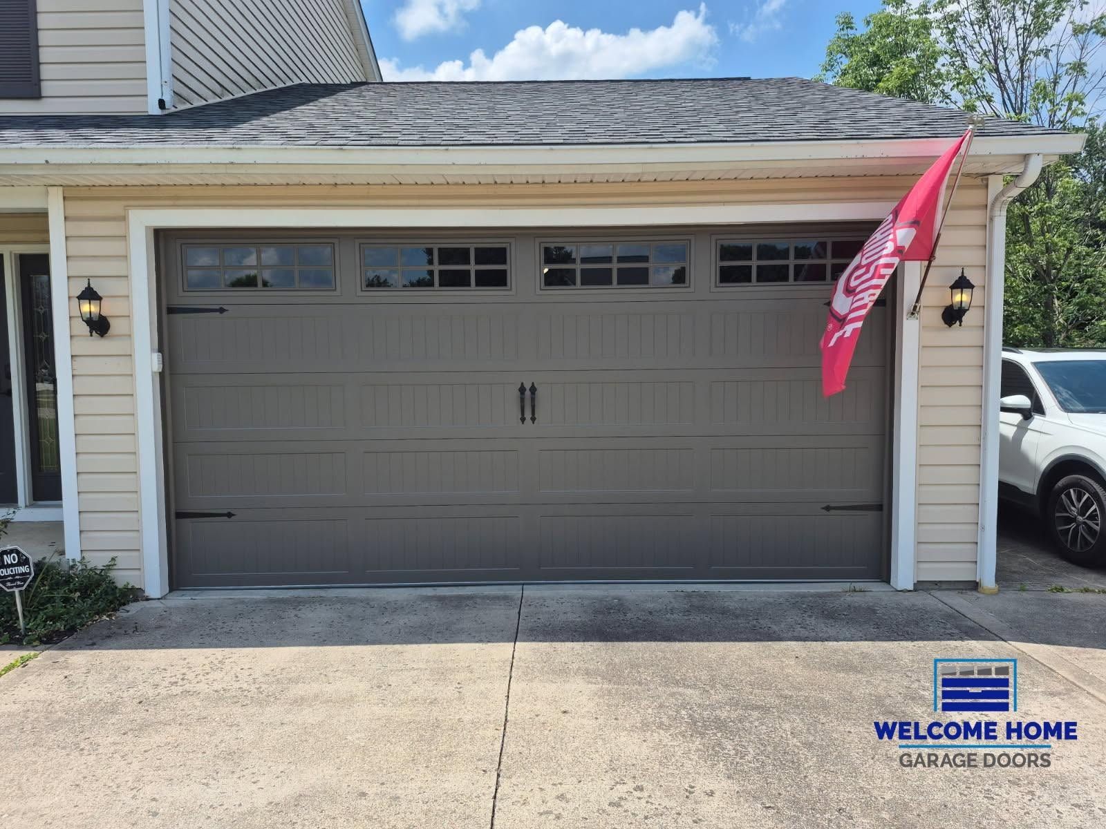 Closed beige garage door with pink flag and small welcome sign beside driveway