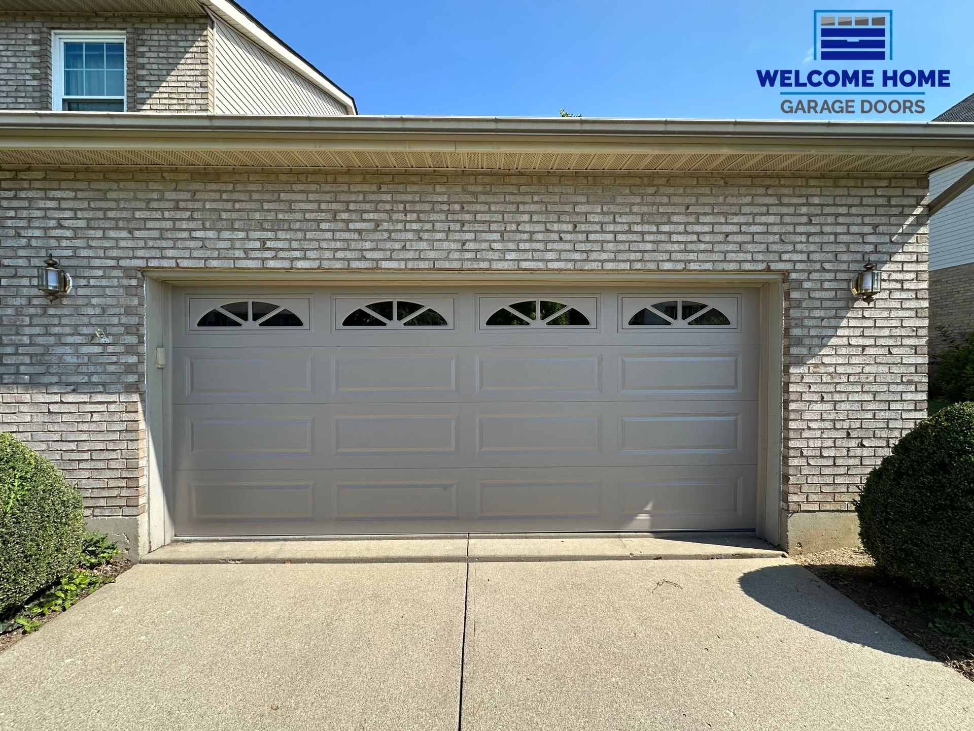 Double garage doors on a stone house with a driveway and “Welcome Home Garage Doors” sign