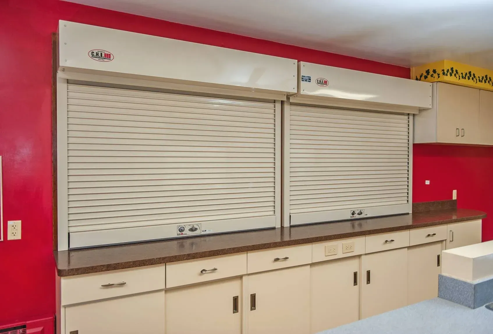 Closed beige roll-down cabinet shutters in a red-and-cream room with countertops and storage cabinets