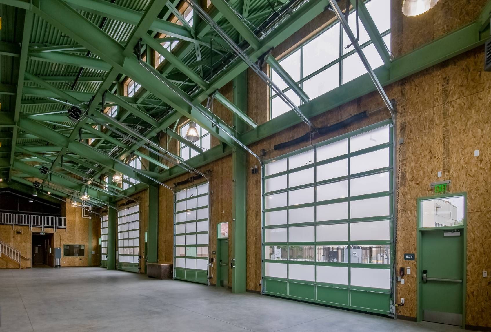 Empty industrial warehouse interior with green steel beams, tall windows, and large garage doors