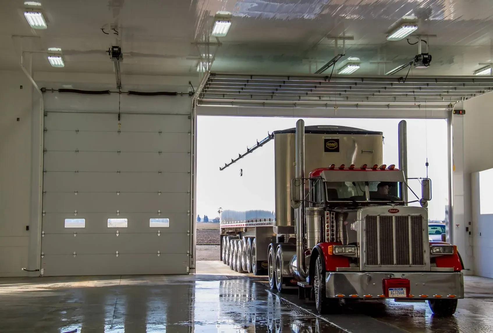 Semi-truck in a wet garage bay beside a large open door, with daylight outside.