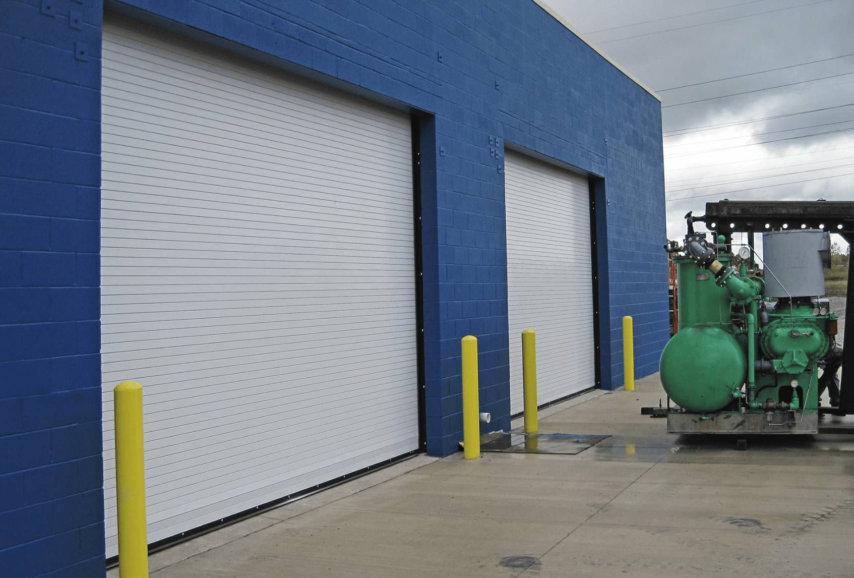 Blue industrial building with white roll-up doors and green machinery beside a wet concrete lot.