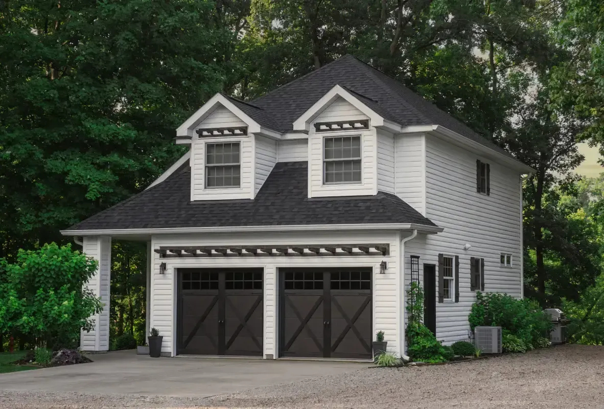 Two-story gray garage with dormer windows, black roof, and double dark doors beside a gravel driveway.