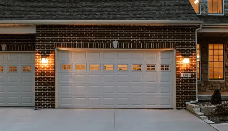 Brick house garage with two white doors, lit by warm exterior lights at dusk