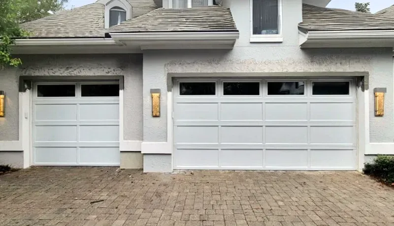 Two white garage doors on a gray house with stone paver driveway and wall lanterns