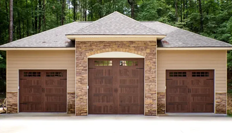 Three-car garage with brown wooden doors and a stone facade beneath a shingled roof