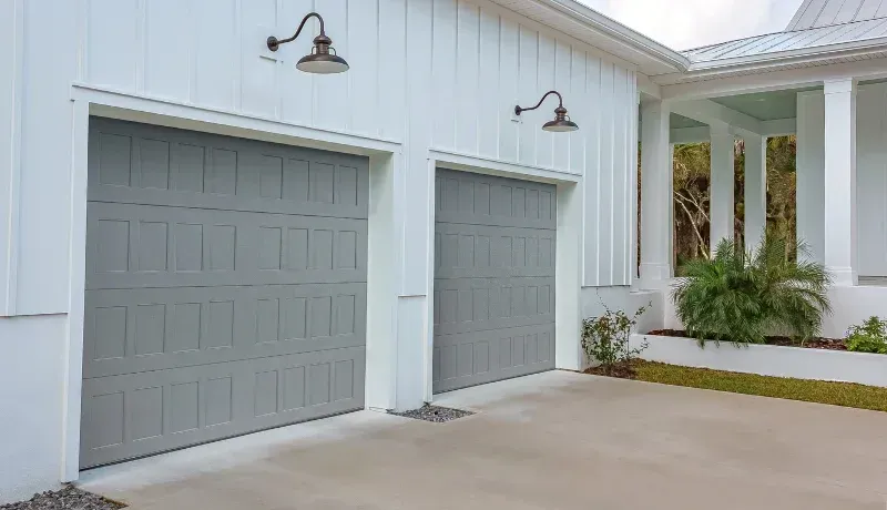 White house exterior with two gray garage doors, wall lights, and a front courtyard with plants.