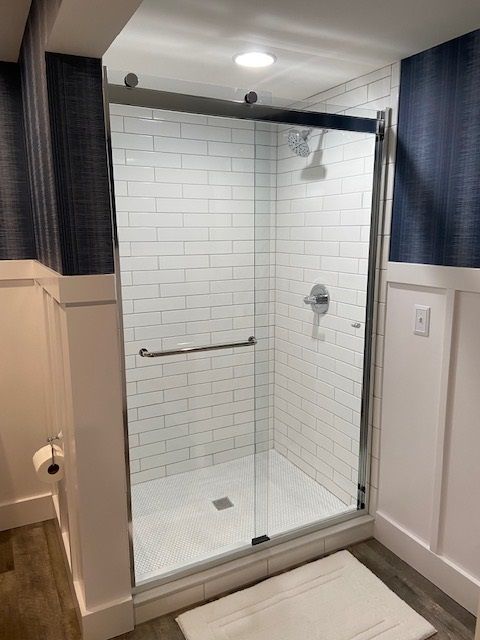 A tiled shower with a glass door in a bathroom with blue wallpaper and white trim.