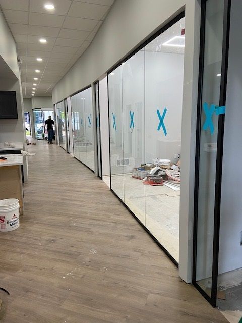 Hallway with glass-walled rooms, light wood floors, blue X symbols on the glass, person walking in the distance.