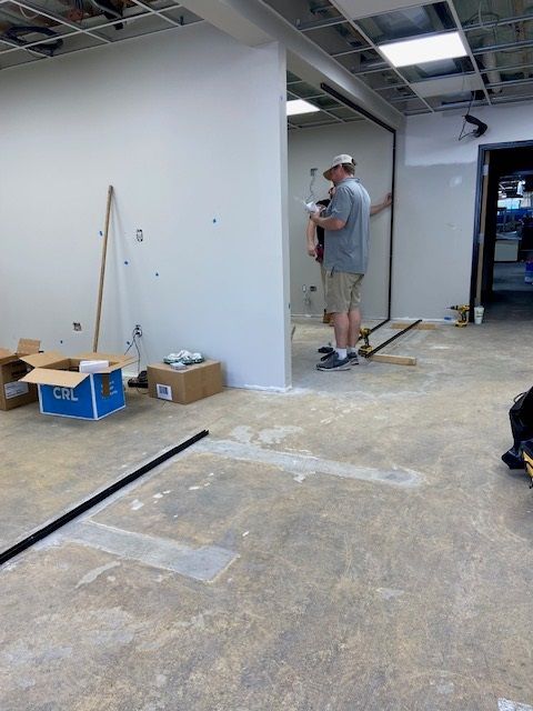 Men working on drywall in a room with a concrete floor. Boxes and tools are present, construction in progress.
