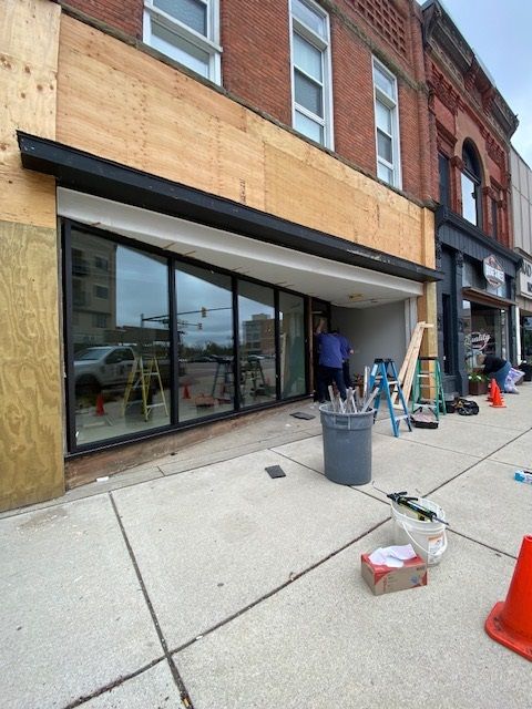Building exterior under construction with large glass windows, black awning. Person, tools, orange cone on sidewalk.