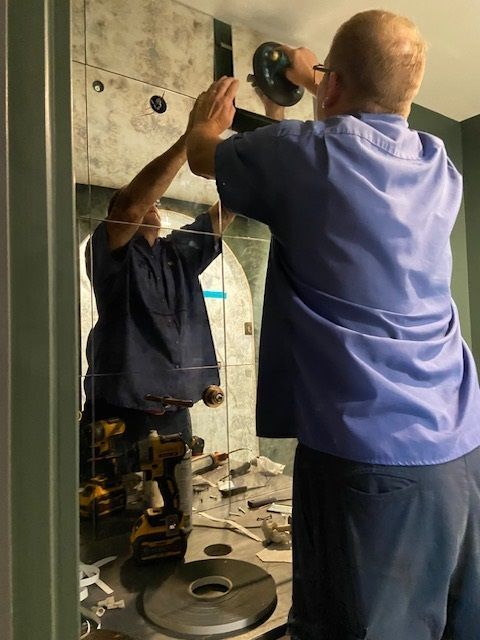 Man in blue shirt installing fixture above a mirror; tools are on the counter.