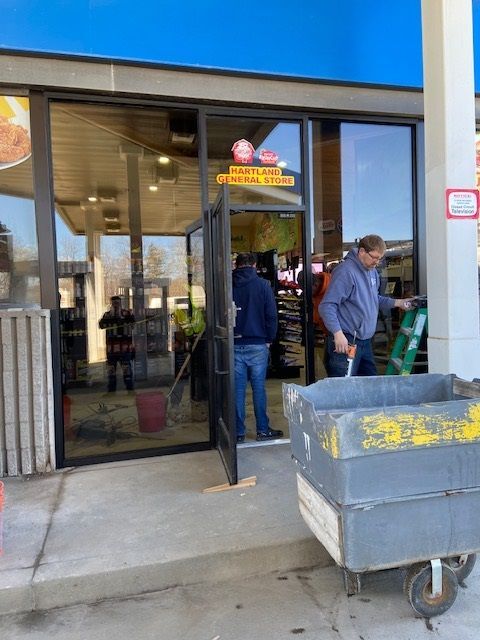 People working on a store entrance. One holds a tool, another stands by the door. A cart is on the right.