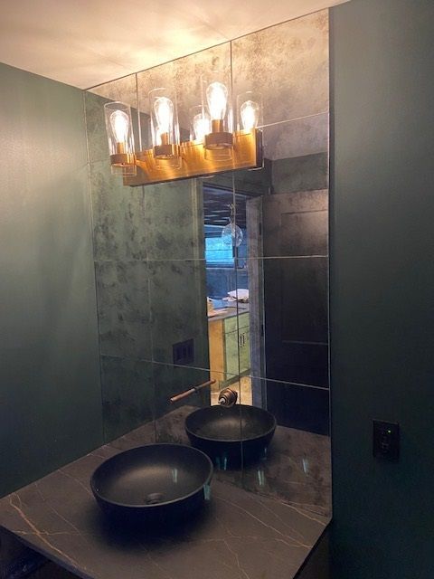 Bathroom with large mirror, black sinks, and gold light fixture. Dark green walls and countertop.